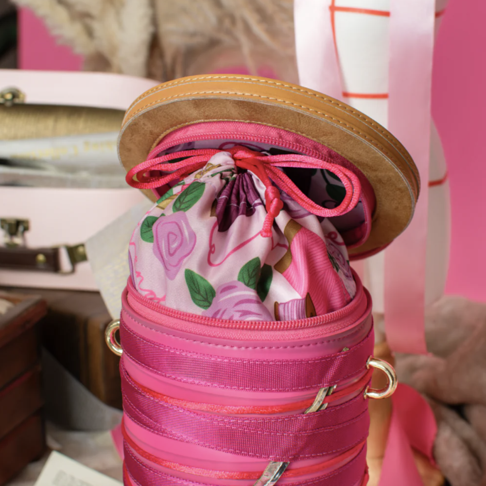 A pink cylindrical zippered bag with a floral-patterned drawstring pouch inside, shown open with a round tan lid resting on top. The bag has pink ribbed fabric bands and gold-tone side rings, photographed in a styled setting with soft pink and neutral background elements.