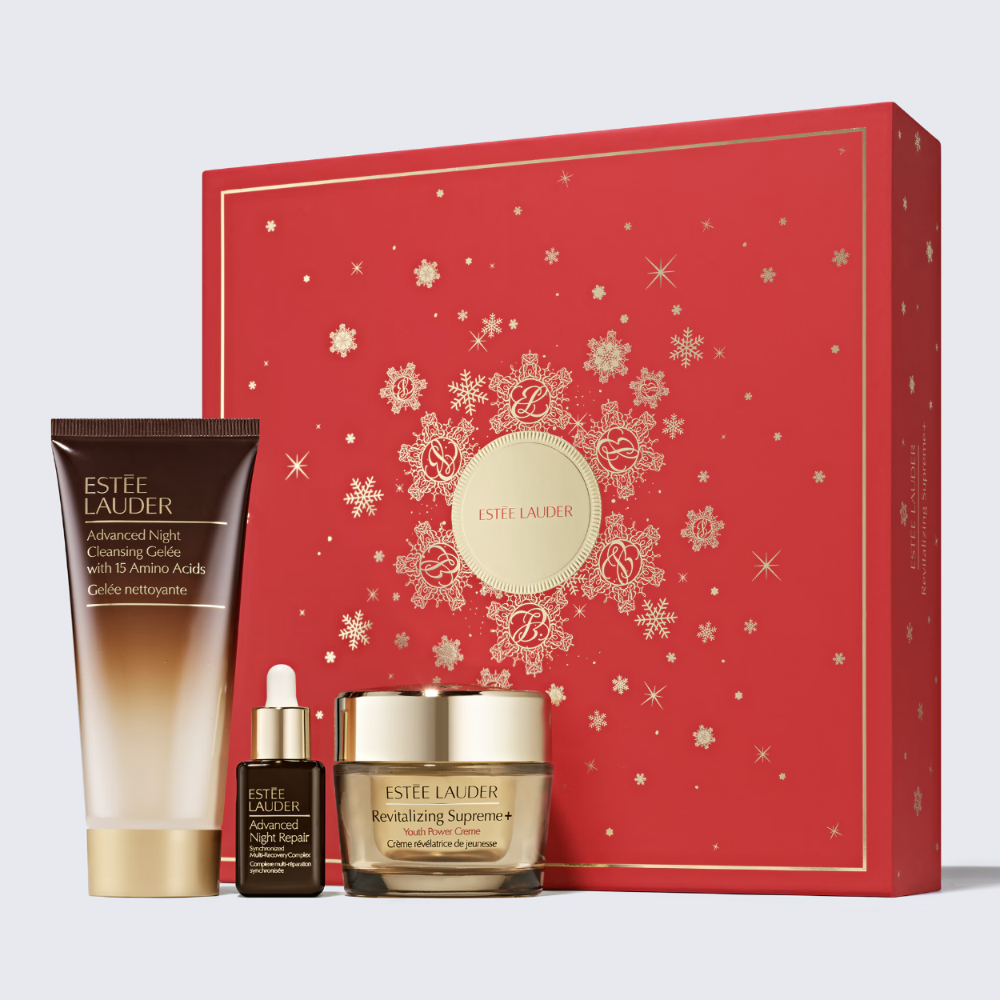 Estée Lauder holiday gift set featuring a red gift box with gold snowflake details, alongside a cream-colored skincare bottle, a small amber dropper bottle, and a gold-lidded cream jar, all arranged on a light gray background.