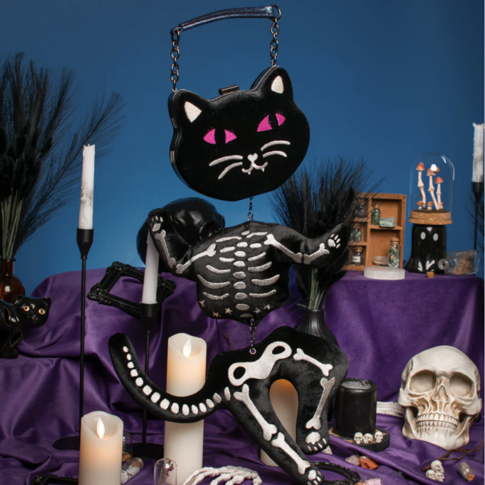 A Halloween-themed tabletop display featuring a black cat-shaped purse with pink eyes, a skeleton cat figurine, candles, a skull, and dark decorative grass against a blue backdrop.