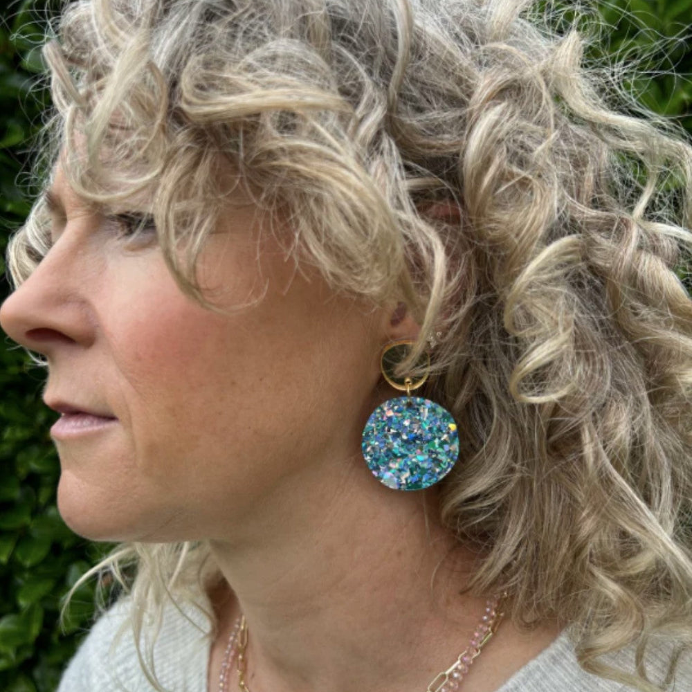 Close-up of a person with curly hair wearing large, colorful earrings against a blurred green background.