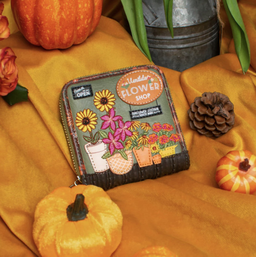 A decorative zip-around wallet styled on a mustard-yellow fabric backdrop with autumn props including pumpkins, a pinecone, flowers, and a metal bucket. The wallet features embroidered flowers, pots, and a sign reading 'Vendula Flower Shop'.