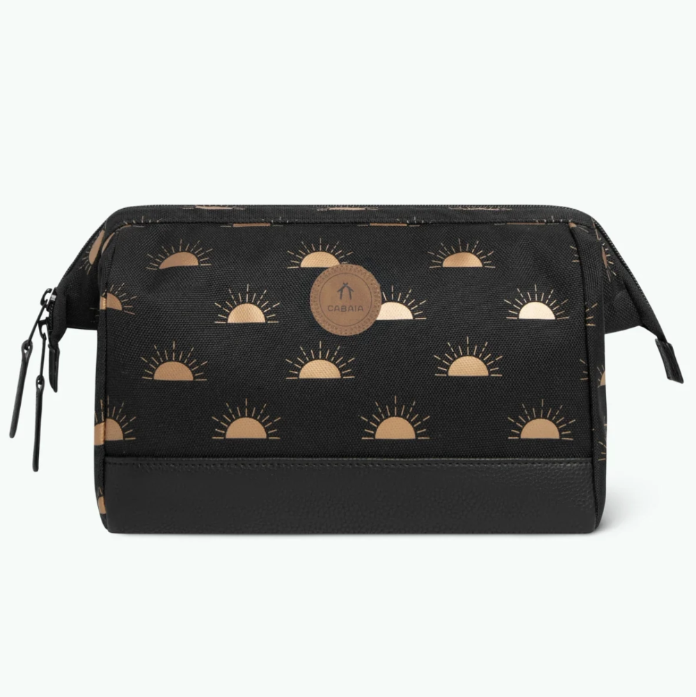 A black cosmetic pouch with a repeating metallic gold sunburst pattern, featuring a circular brown leather brand patch in the center and a black textured bottom panel.
