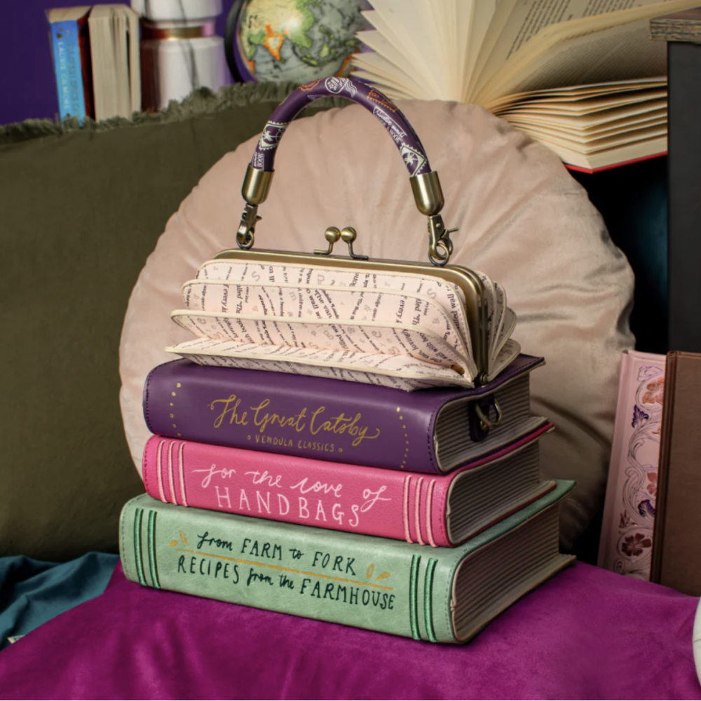 A bag in the shape of a stack of four decorative books on a purple fabric surface, topped with a small kiss-lock clutch bag featuring a purple patterned handle and cream patterned body. The books include titles such as 'The Great Catsby', 'In the Love of Handbags', and 'From Farm to Fork'.