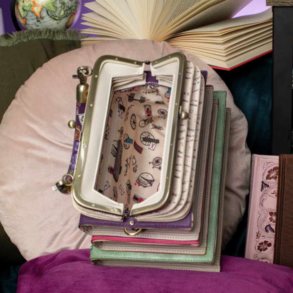 A stack of vintage-style book covers and journals with a matching open clutch bag resting on top, set against a cozy background with an open book, cushions, and a purple fabric surface.