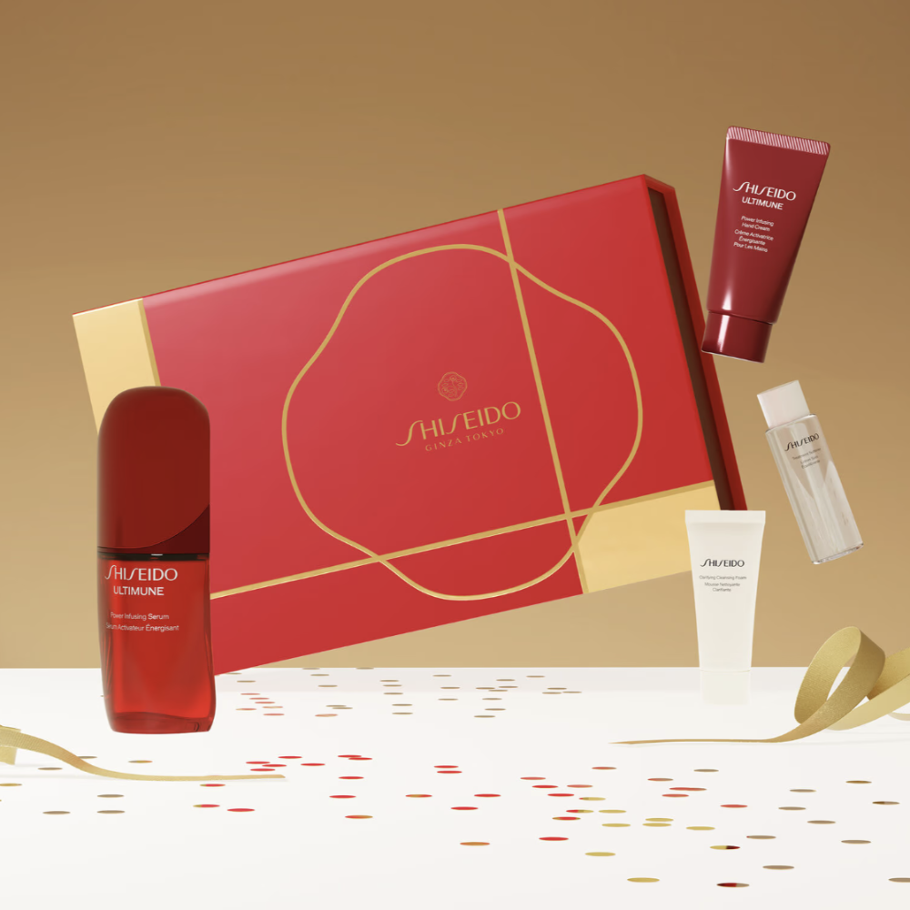 Shiseido gift set featuring a large red box with gold detailing, a red serum bottle, a red hand cream tube, and a small clear bottle, arranged on a white surface with gold ribbons and confetti against a warm beige background.