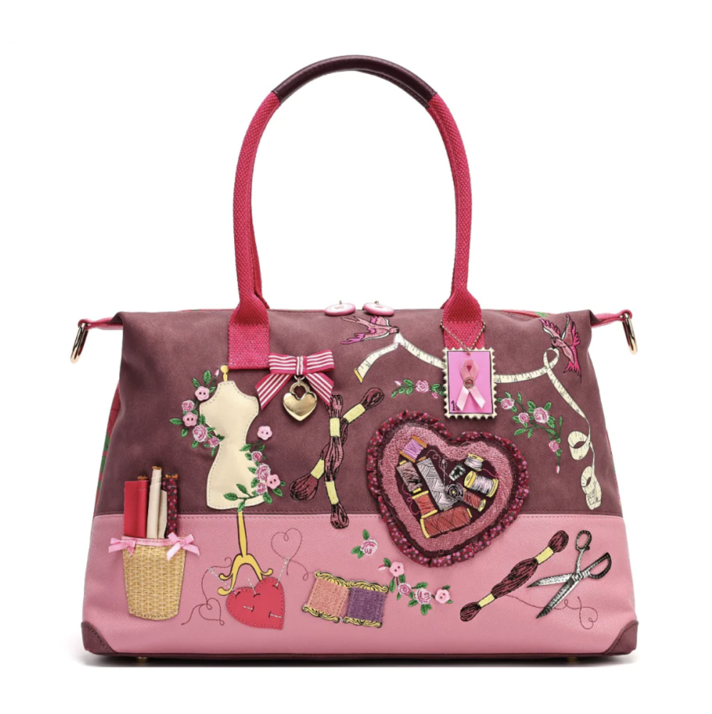 A pink and mauve handbag decorated with sewing-themed appliqués, including a mannequin, scissors, thread, lipstick hearts, and a pink ribbon charm, shown on a plain white background.