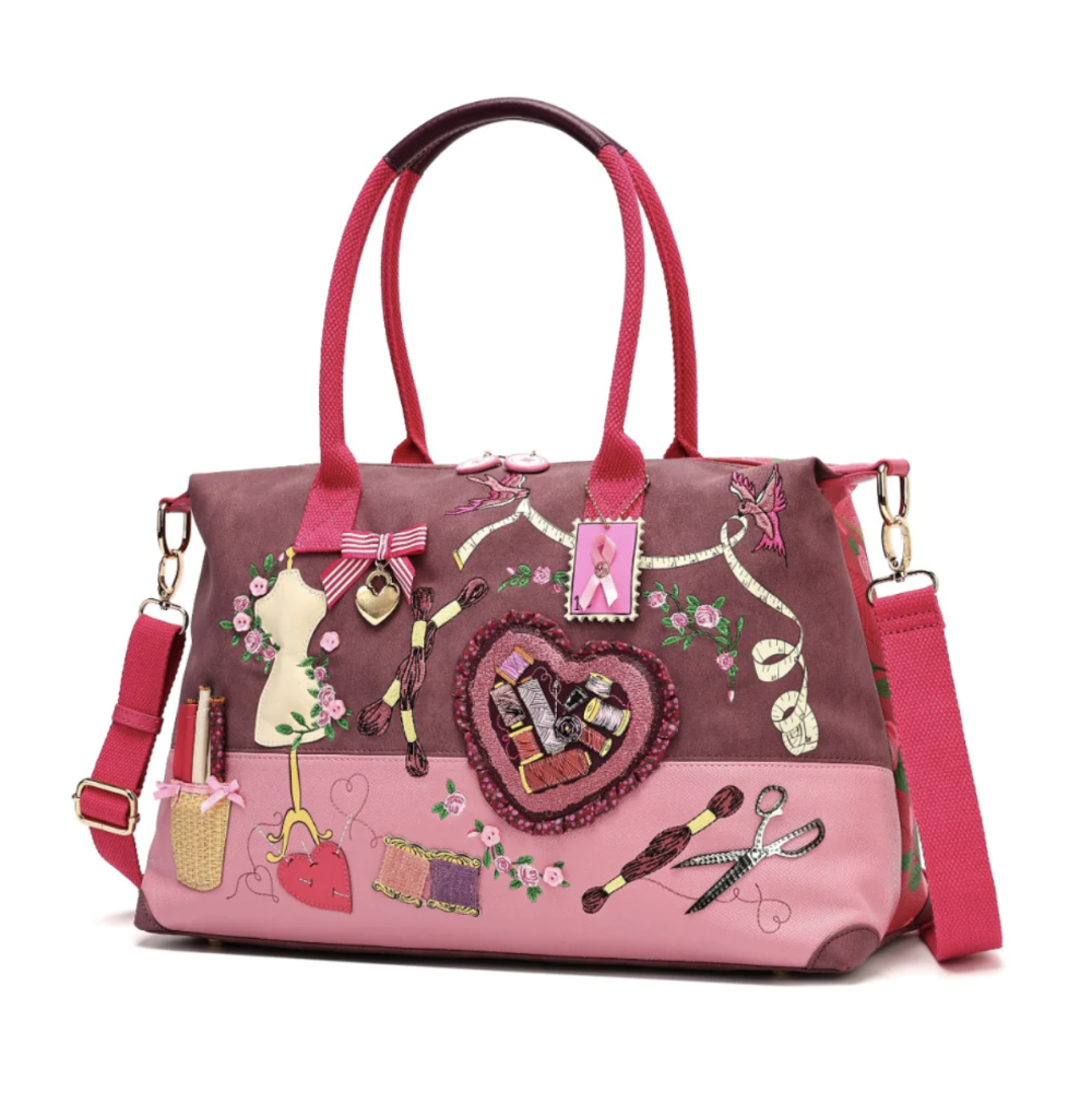 A pink and mauve sewing-themed tote bag with embroidered details, dual top handles, a detachable shoulder strap, and decorative patches including a mannequin, scissors, thread, and a heart-shaped patch.