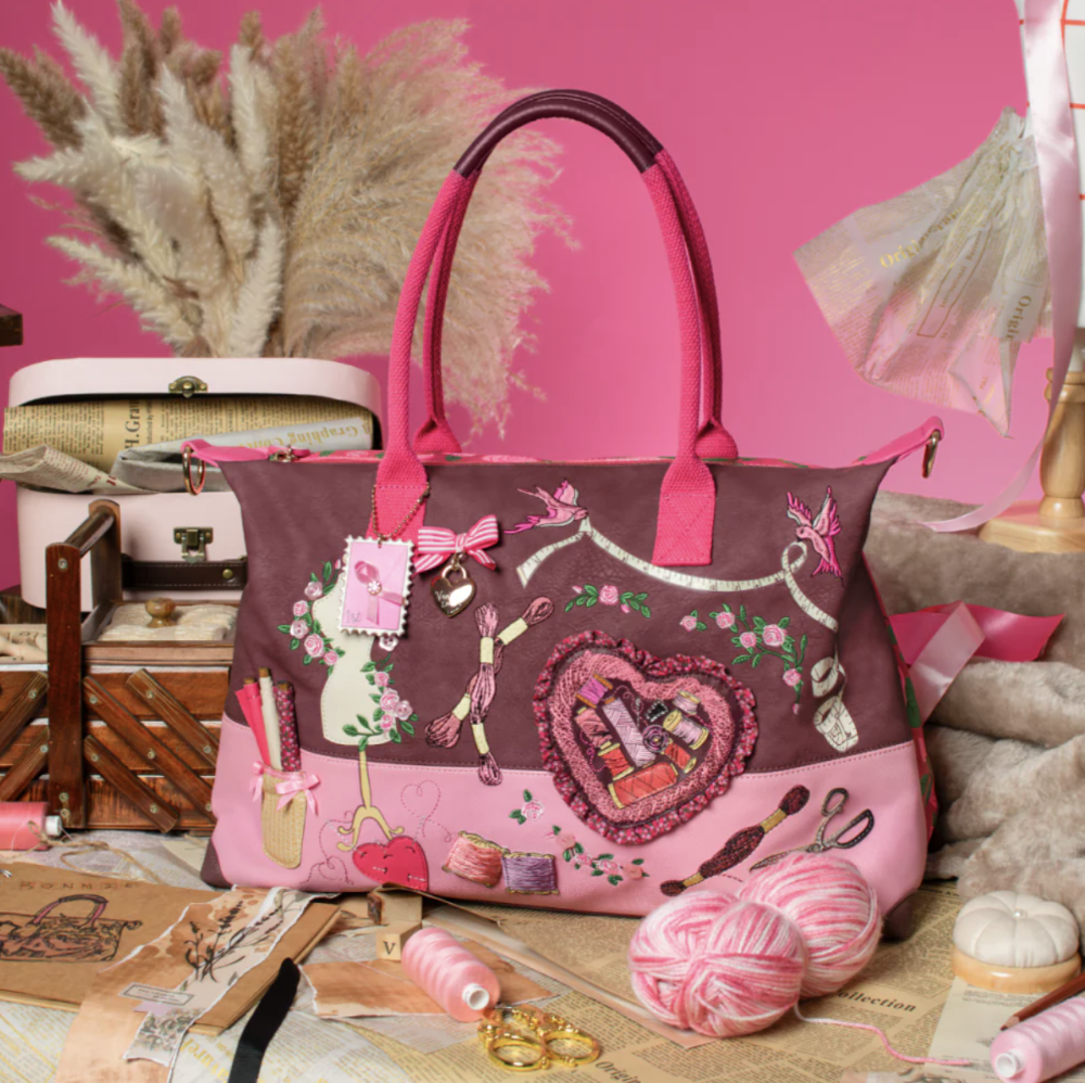 A pink and mauve patchwork tote bag with embroidered sewing and heart motifs, styled on a table with yarn, spools of thread, and sewing notions against a pink background.