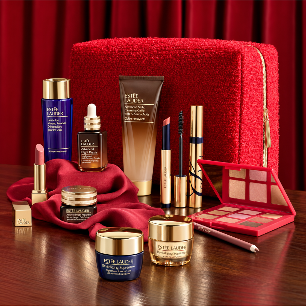 A luxury beauty gift set arranged on a dark wooden table against a deep red curtain backdrop. The set includes Estée Lauder products such as the Advanced Night Repair serum, and Advanced Night Repair Eye Gel and more