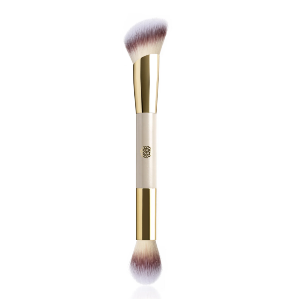 A dual-ended makeup brush with a cream-colored handle, gold ferrules, and soft bristles that fade from beige to light purple, shown on a clean white background.