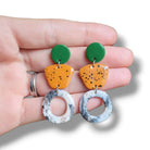 Hand holding colorful earrings with green, orange, and gray components on a white background