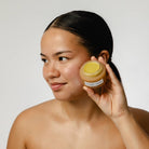 Woman holding a jar of cream to her face against a plain background