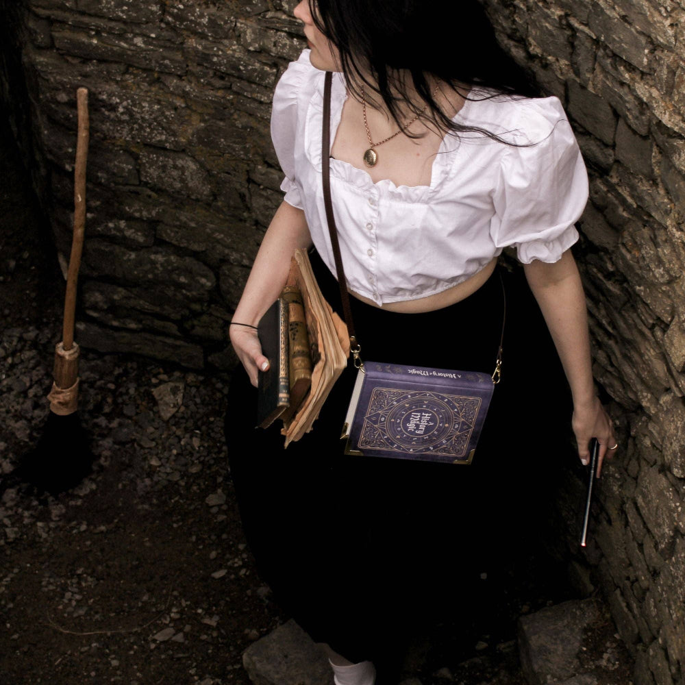 Person in a white shirt and black skirt holding books and a broomstick in a dark alley.