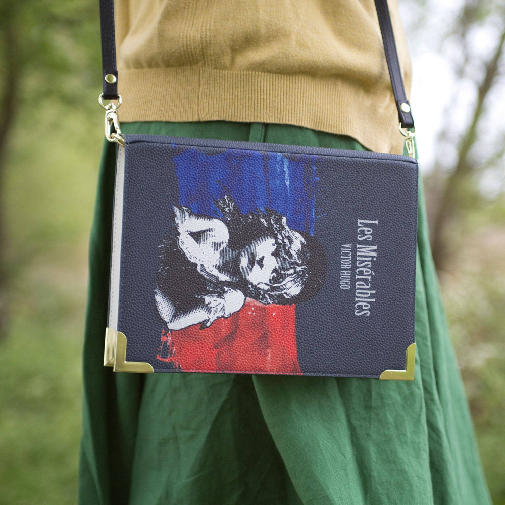 Handbag with 'Les Miserables' design held by a person wearing green pants and a beige sweater.