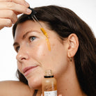 Woman applying face oil to her forehead with a dropper, holding a bottle labeled 'UpCircle Face Oil'.
