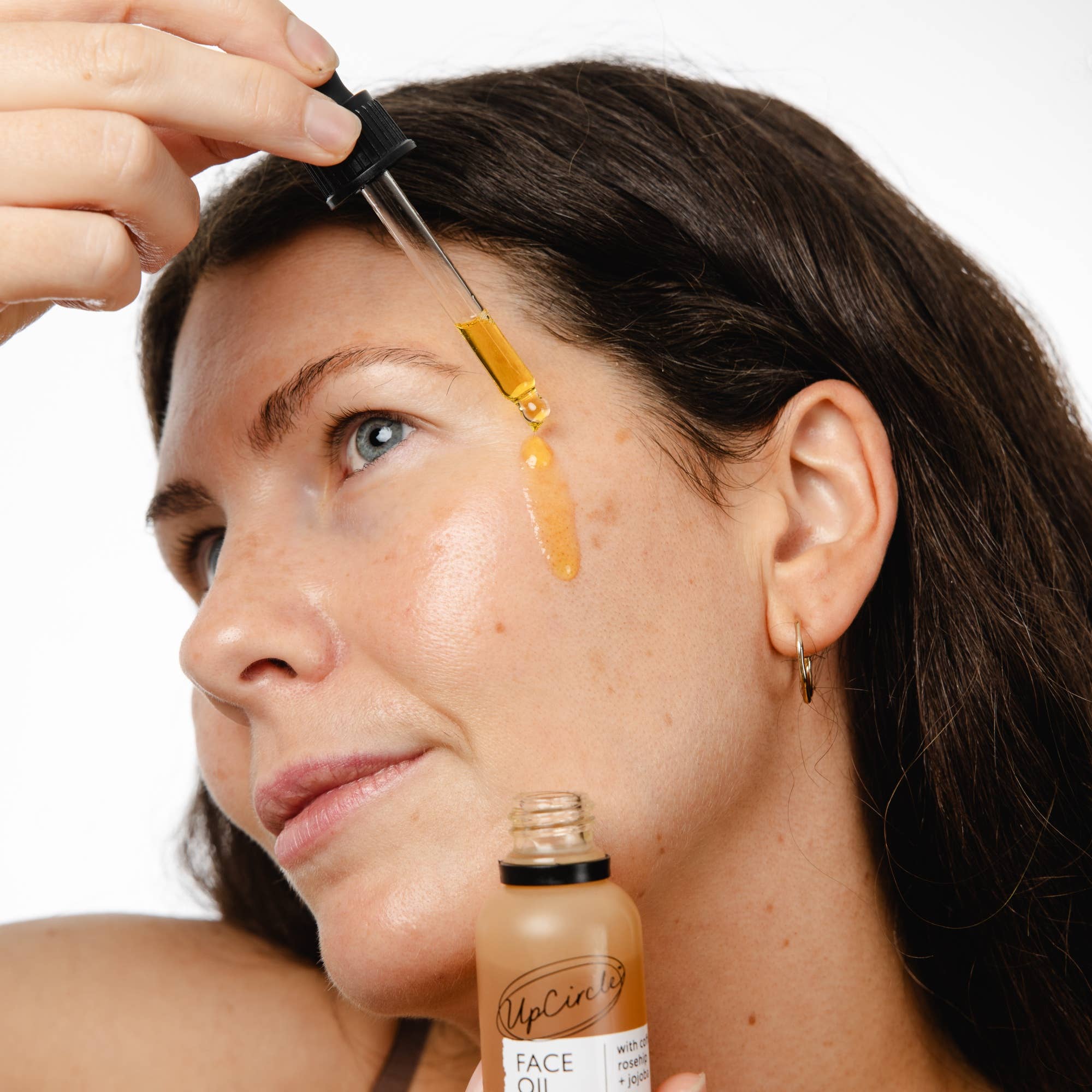 Woman applying face oil to her forehead with a dropper, holding a bottle labeled 'UpCircle Face Oil'.