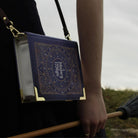Book-shaped handbag with 'History of Magic' design held by a person outdoors.