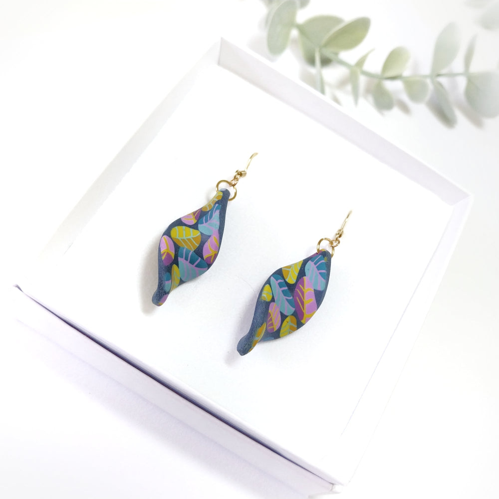 Colorful leaf-shaped earrings on a white surface with a plant branch in the background