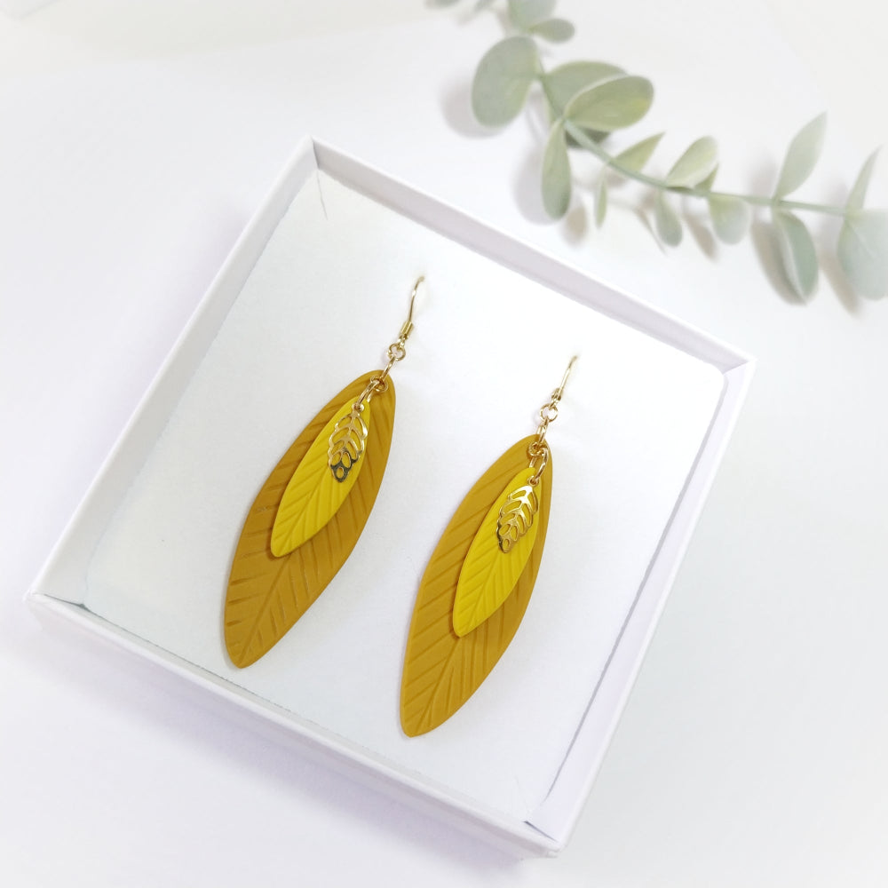 Yellow leaf-shaped earrings on a white box with green leaves in the background