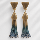 A pair of statement dangle earrings with textured gold-tone metal tops and teardrop-shaped links, featuring braided tassels that transition from gold at the top to blue at the bottom, set against a light gray background with soft shadows.