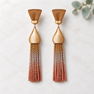 A pair of statement dangle earrings with textured gold-tone metal tops and long braided tassels that fade from gold to copper and rose-gold, displayed on a light marble surface with a small green leaf in the corner.