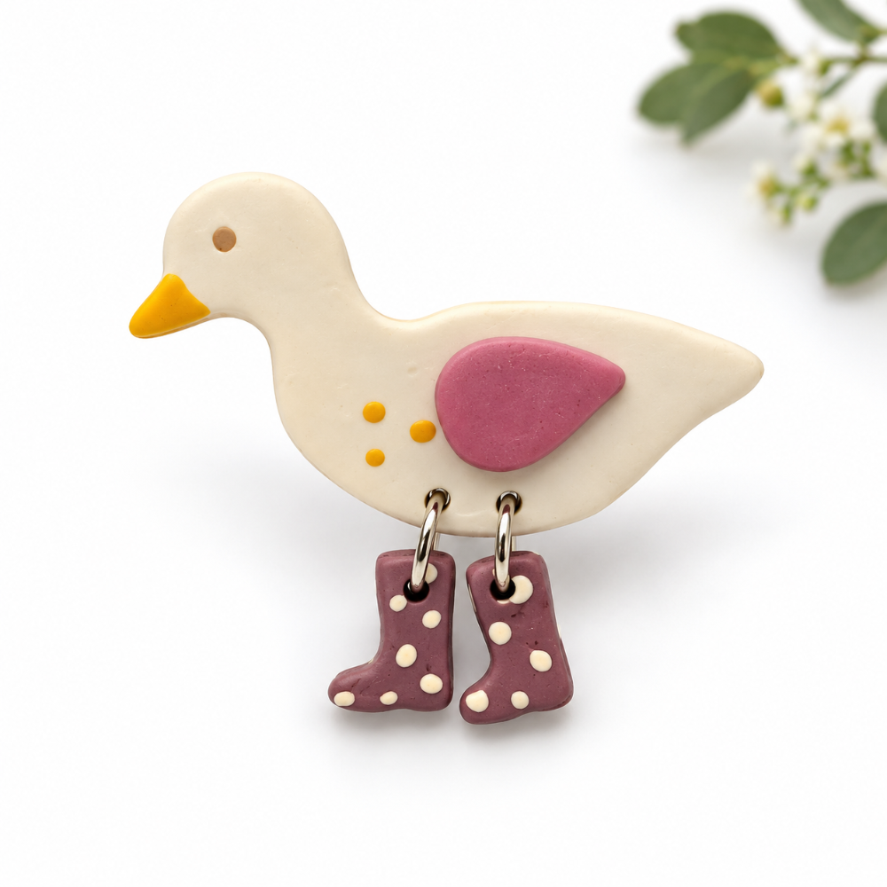 A duck-shaped jewelry brooch pin piece with a cream body, yellow beak, pink wing, and dangling boot charms in mauve with cream polka dots, shown on a white background with soft green leaves in the corner.