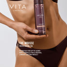 Person holding a bottle of Vita Liberata The Mousse with product details on a light gray background