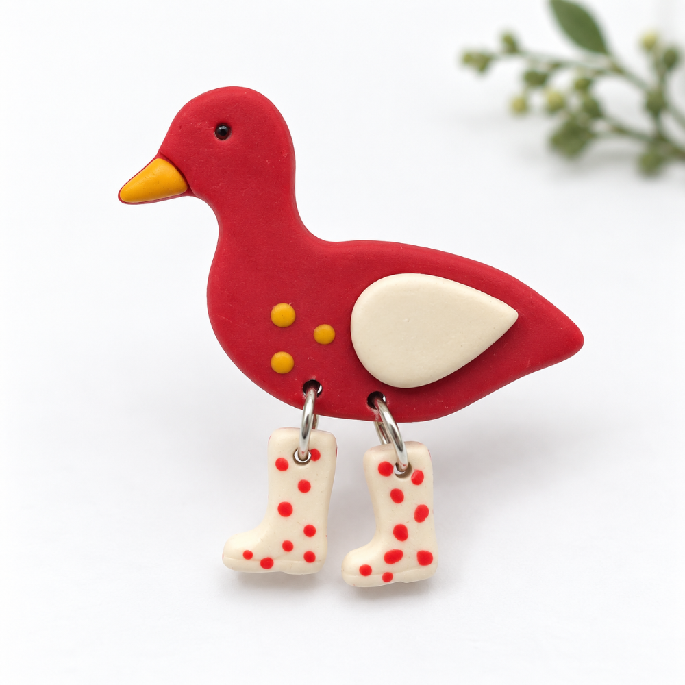 A red duck-shaped brooch pin with a yellow beak, cream wing, and two dangling cream rain boots with red polka dots, shown on a white background with a soft green plant in the upper right.
