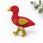 A red bird-shaped brooch pin with a yellow beak, yellow wing, and yellow-green spotted legs, photographed on a clean white background with a soft leafy blur in the corner.