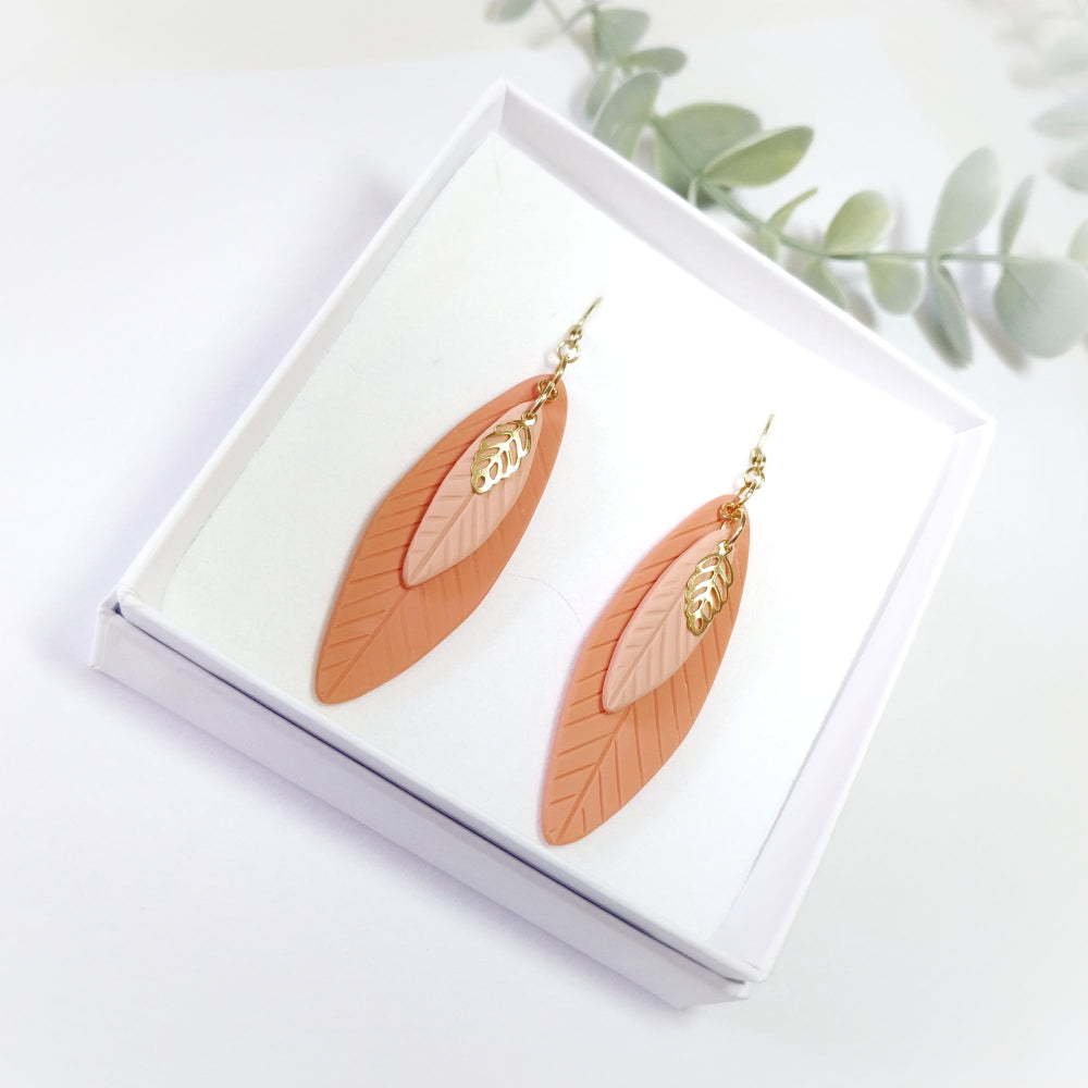 Pair of leaf-shaped earrings on a white box with greenery in the background