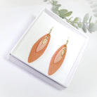 Pair of leaf-shaped earrings on a white box with greenery in the background