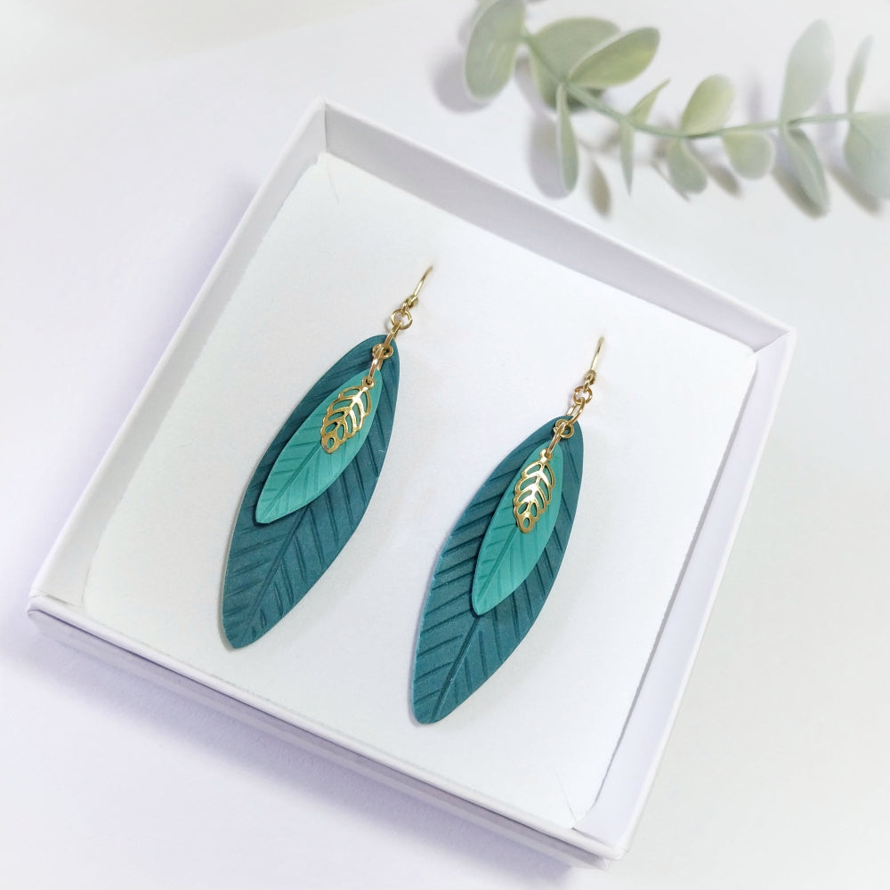 Teal feather-shaped earrings with gold accents on a white box with a plant leaf in the background.