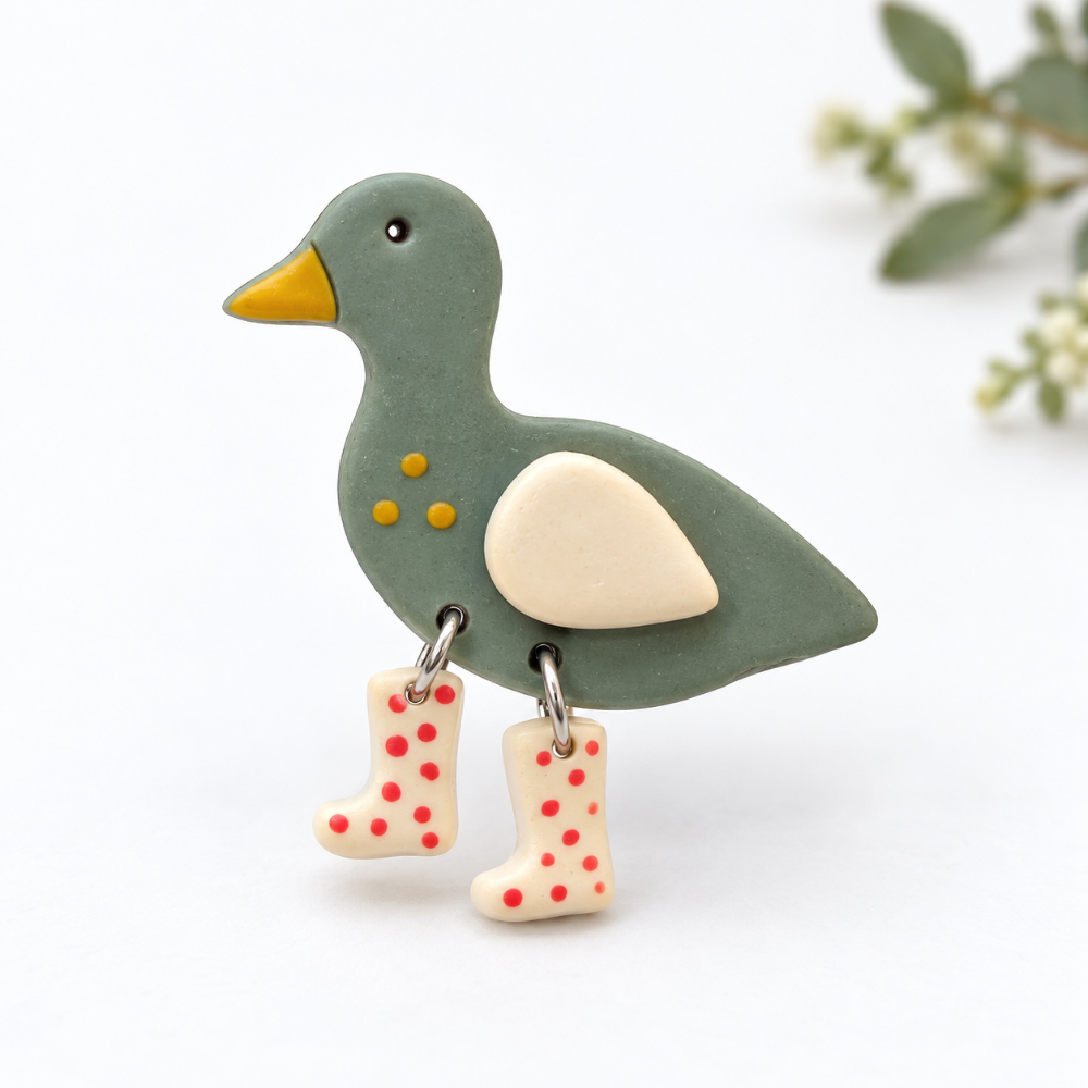 A cute duck-shaped brooch or pin in muted sage green with a yellow beak, cream wing, and small yellow spots, with two dangling cream rain boots decorated with red polka dots.
