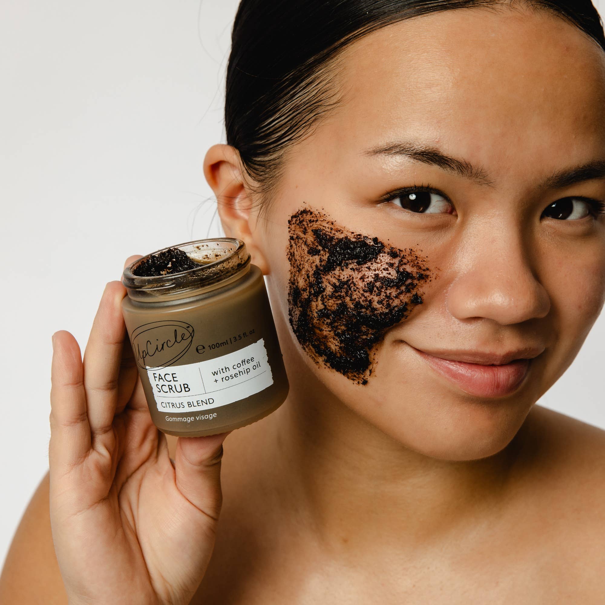 Woman applying coffee scrub to her face with a jar of scrub in hand.