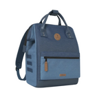 A blue canvas backpack with a dark navy base, featuring a front zippered pocket, top handles, and a circular tan leather logo patch on a light background.
