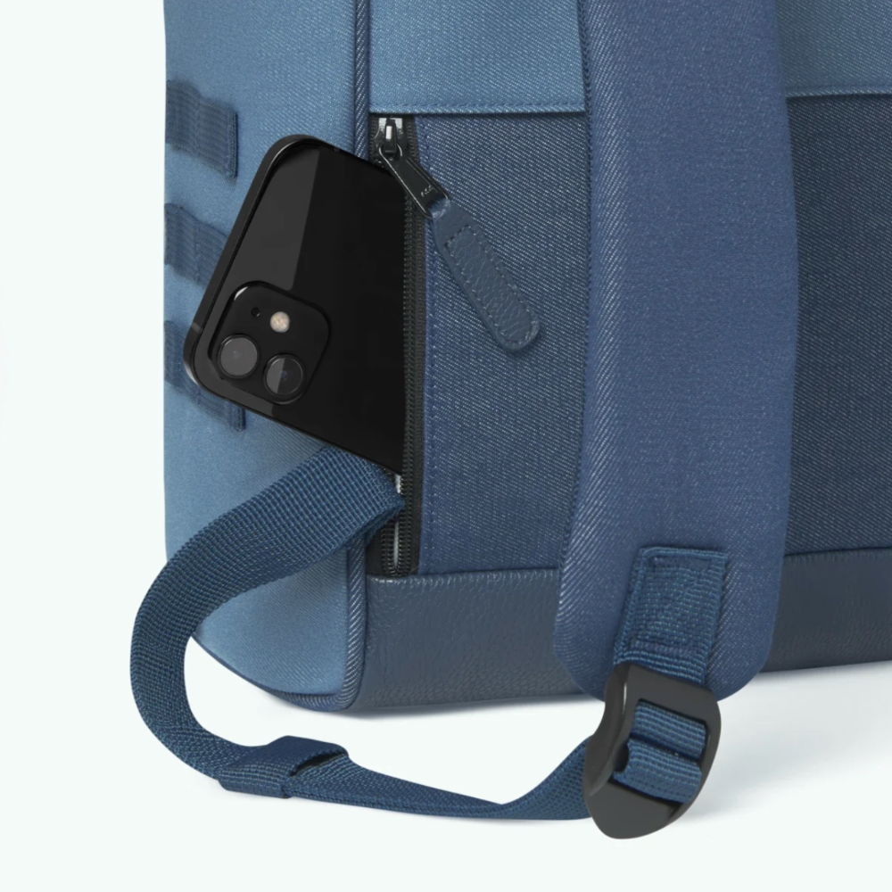 Close-up of a blue backpack showing a side pocket with a black smartphone tucked inside, along with a blue shoulder strap and zipper detail on a clean white background.
