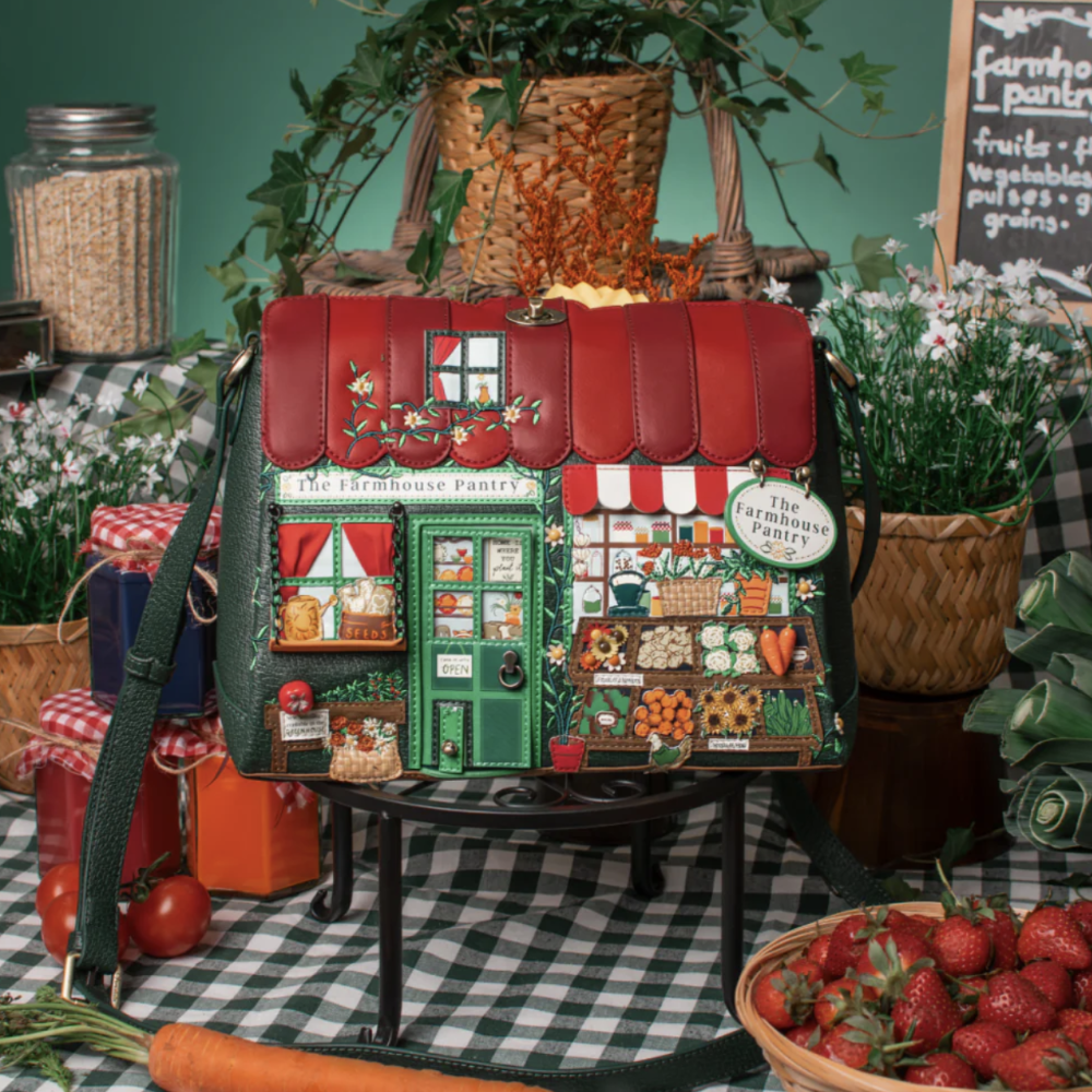 A decorative shoulder bag shaped like a small farmhouse storefront sits on a table in a styled rustic setting. The bag has a red roof, green walls, and illustrated market stalls with vegetables, fruit, and a sign reading 'The Farmhouse Store.