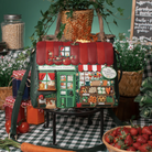 A decorative shoulder bag shaped like a small farmhouse storefront sits on a table in a styled rustic setting. The bag has a red roof, green walls, and illustrated market stalls with vegetables, fruit, and a sign reading 'The Farmhouse Store.