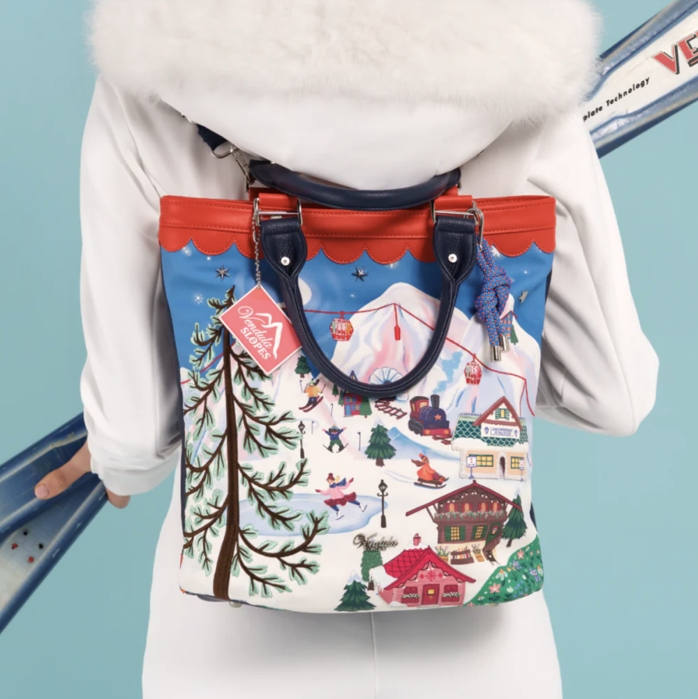 A person wearing a white coat with a fluffy hood, shown from the back while wearing a colorful ski resort backpack. The backpack features a detailed winter mountain scene with cabins, trees, and skiers, accented with red trim and dark blue handles.