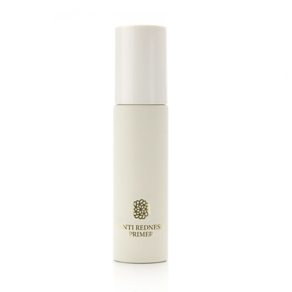 A cylindrical cosmetic bottle with a white cap and a cream-colored body, featuring the text 'ANTI REDNESS PRIMER' and a small gold floral emblem, set against a plain white background.