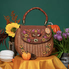 A brown wicker handbag with a braided top handle, decorated with colorful floral embroidery and appliques on the front flap. The bag is styled on a yellow fabric surface with autumn props including pumpkins, tulips, and a sunflower against a dark green background.