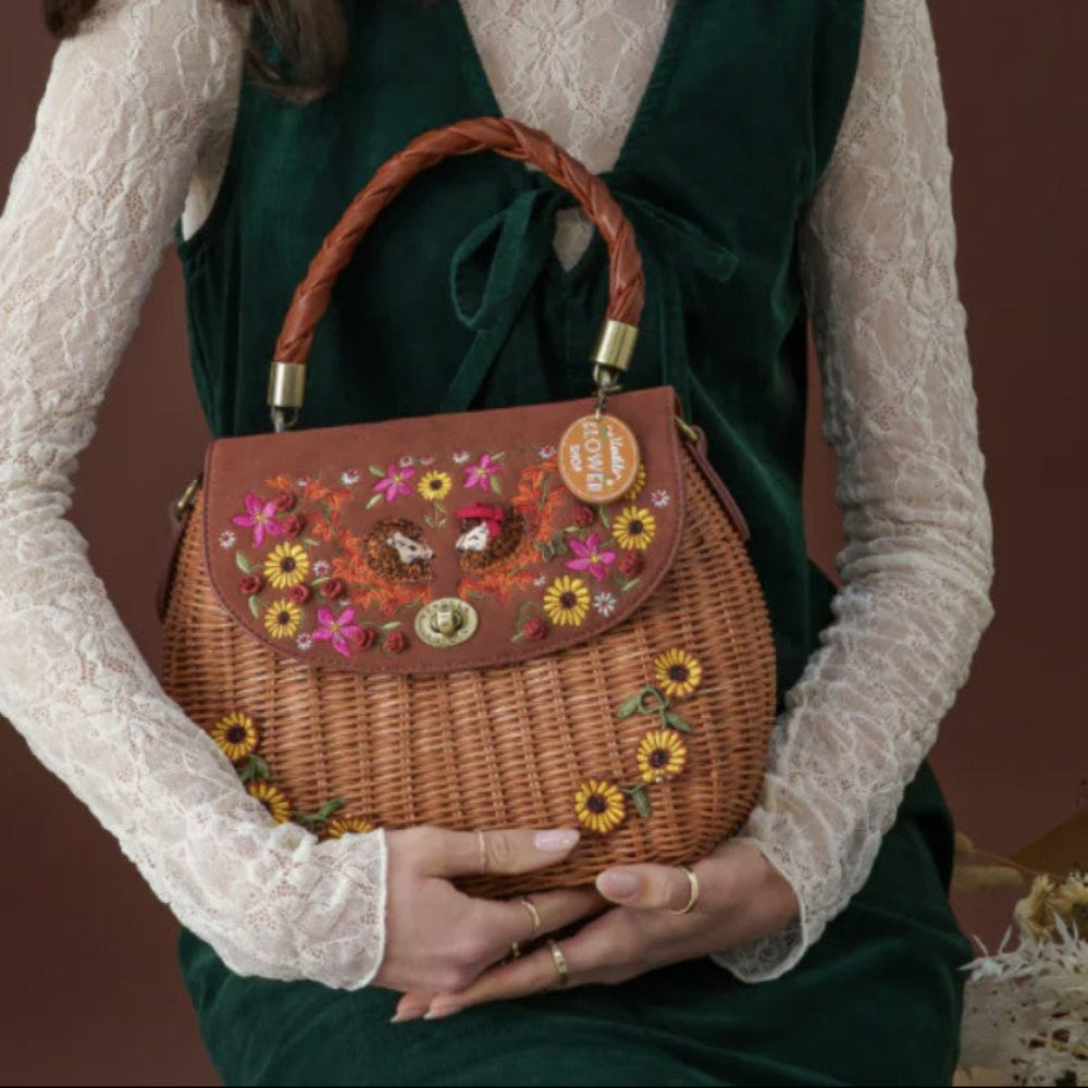 A woman in a dark green velvet dress and white lace top holds a brown woven wicker handbag with floral embroidery, sunflower details, and a braided top handle against a dark brown background.