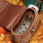 A brown woven wicker handbag with a brown leather interior flap, shown open on a golden satin fabric with autumn props like a pumpkin. 