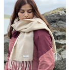 A woman wearing a cream-colored woven scarf with fringe and a small white label that reads 'merlin & co' is pictured outdoors against a rocky, overcast background.