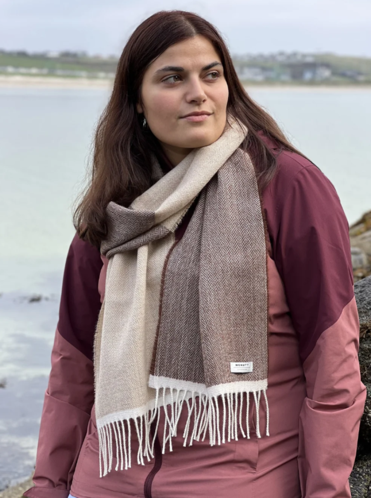 A woman wearing a large woven scarf in beige and brown with white fringe, styled over a dusty rose jacket, standing outdoors near the water.
