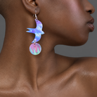 A close-up of a model wearing a pair of dangling earrings shaped like a blue and purple bird with a pink and iridescent circular charm below, set against a neutral gray background.