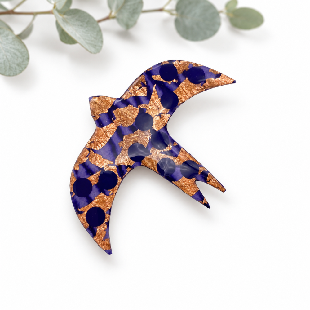 A bird-shaped decorative brooch or pin made from patterned fabric, decorated with oblackcurrant and gold circular embellishments, photographed on a white background with eucalyptus leaves at the top.