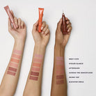 Three hands holding makeup products with color swatches on arms against a white background