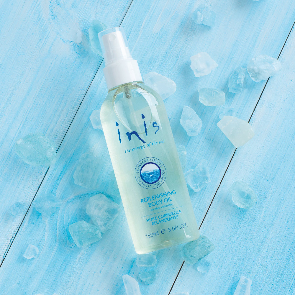 Bottle of Inis replenishing body oil on a blue wooden surface with ice cubes.
