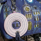 A close-up of a blue fabric bag featuring an illustrated clock tower and moon design, with a pink circular ring charm attached by silver metal hardware and a dark blue ribbon.